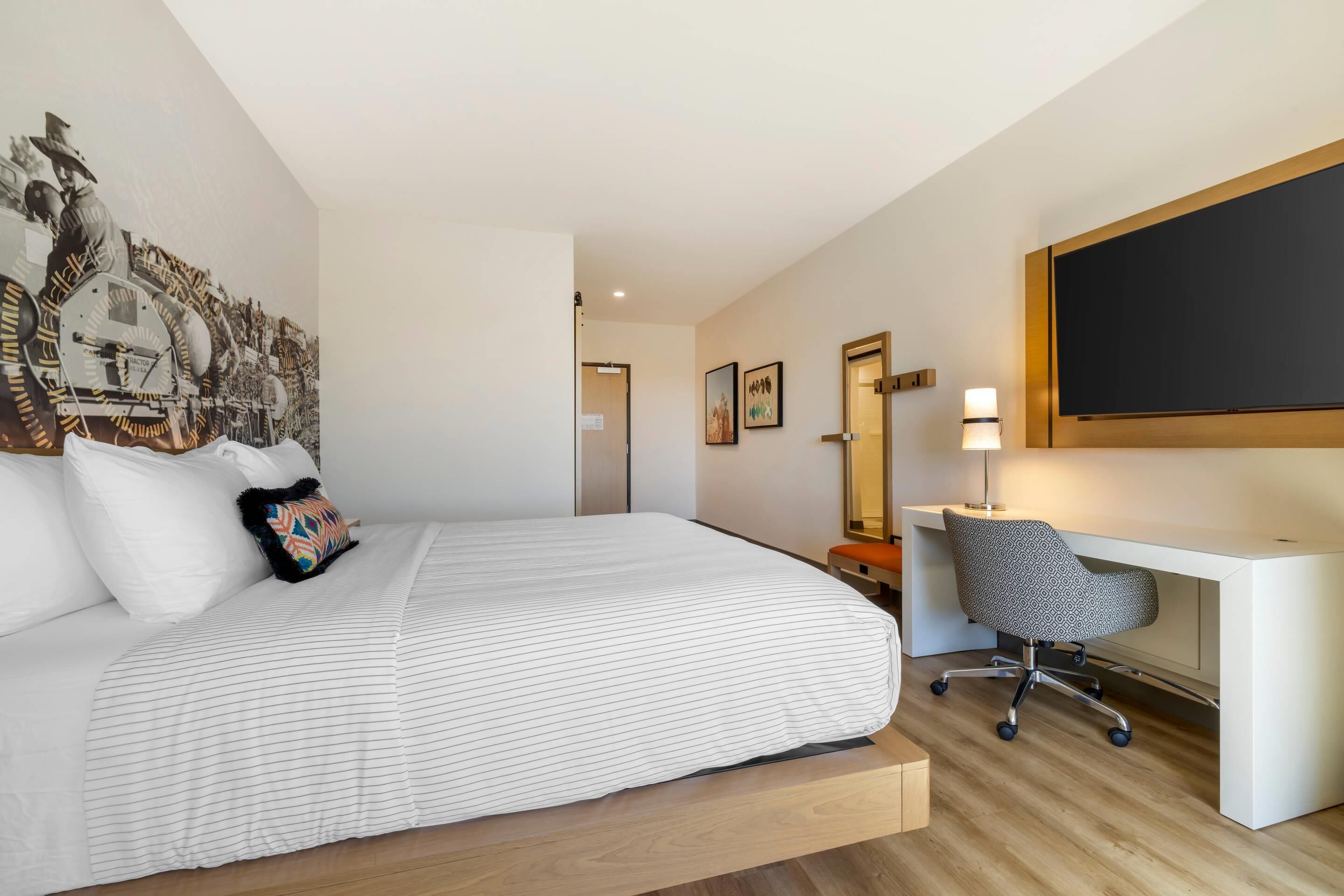 Habitación de hotel con cama, escritorio, silla, lámpara de escritorio y un cuadro grande en la pared.