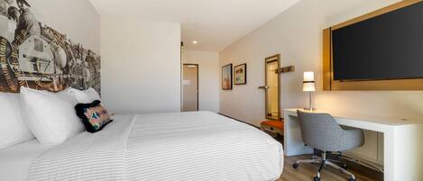 Habitación de hotel con cama, escritorio, silla, lámpara de escritorio y un cuadro grande en la pared.