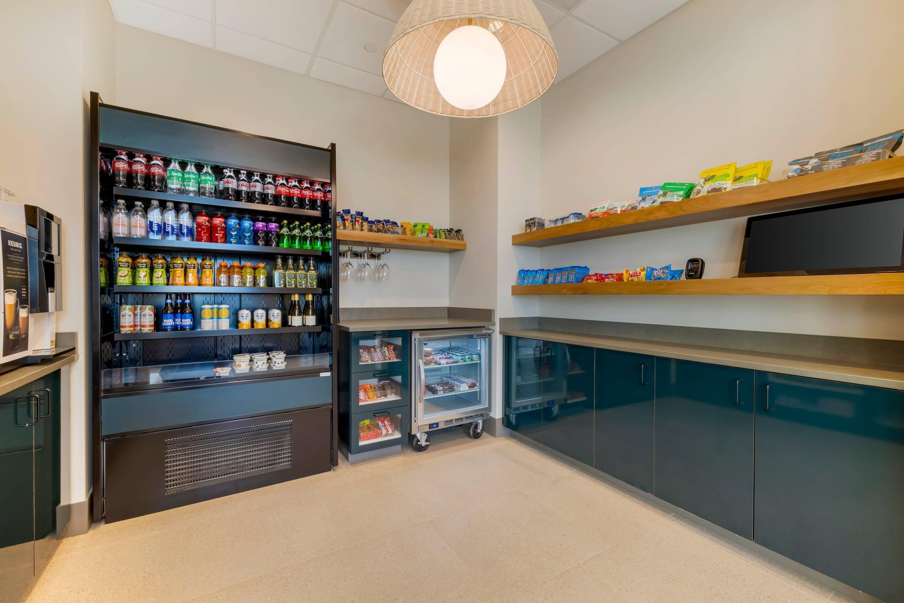 Una habitación bien iluminada con un dispensador de bebidas repleto de diversas opciones, un refrigerador pequeño y estantes con aperitivos y condimentos.