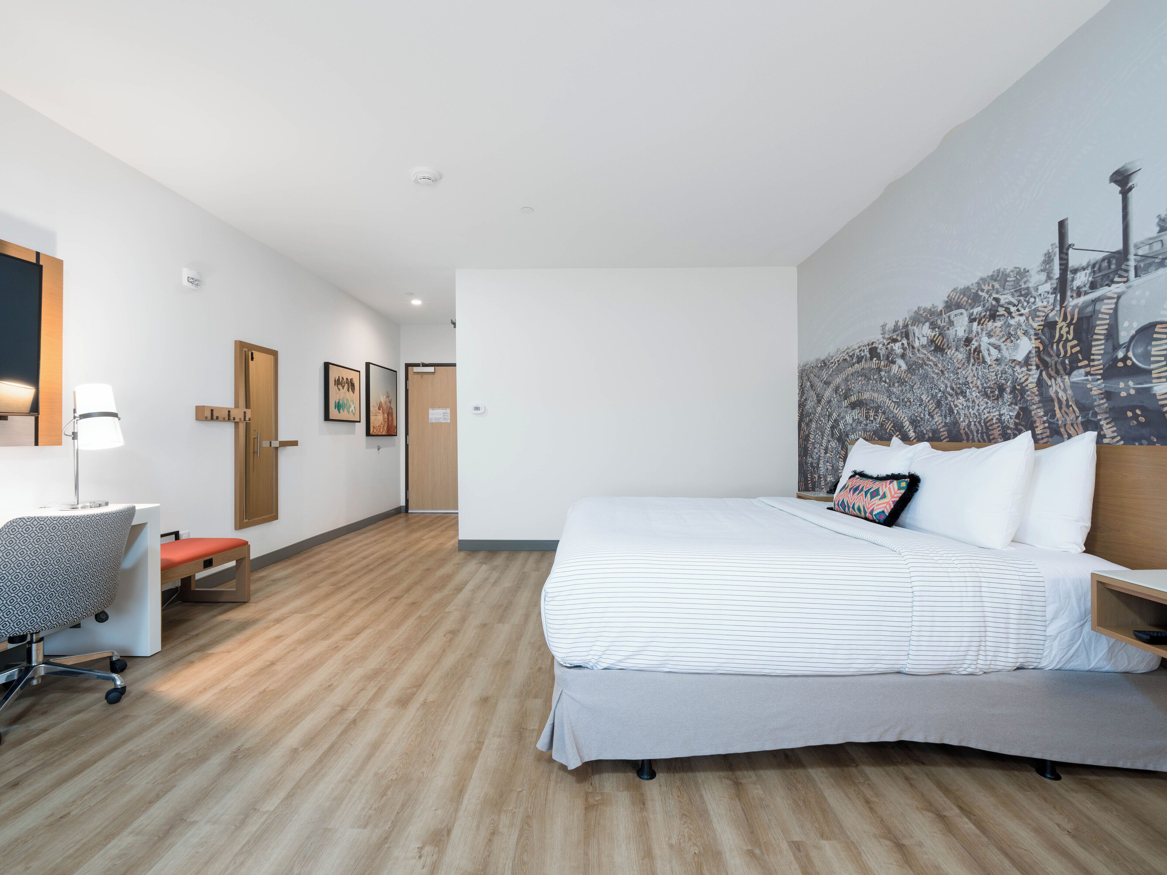 Una habitación de hotel moderna con una cama grande, un escritorio con silla, piso de madera y una pared con una pintura de paisaje urbano.