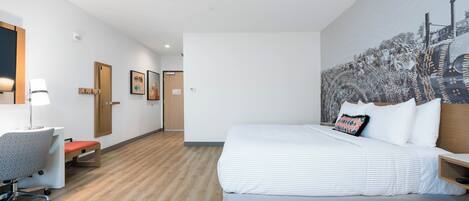 Una habitación de hotel moderna con una cama grande, un escritorio con silla, piso de madera y una pared con una pintura de paisaje urbano.