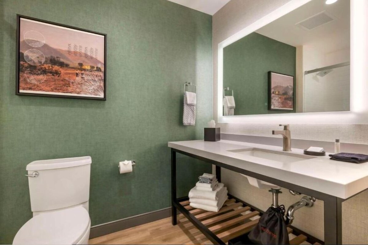 Un baño con un inodoro blanco, un lavamanos con espejo y un cuadro en la pared.