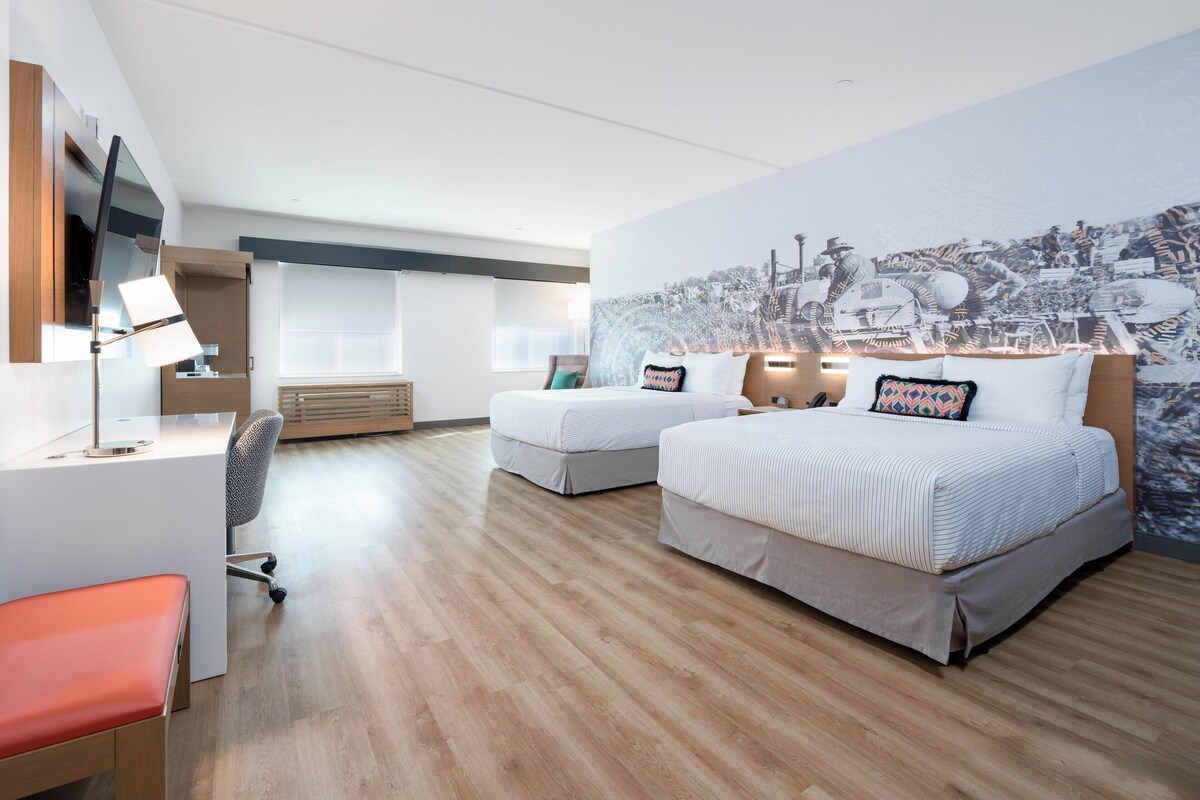 A modern hotel room with two beds, a desk, a chair, and a large mural on the wall.