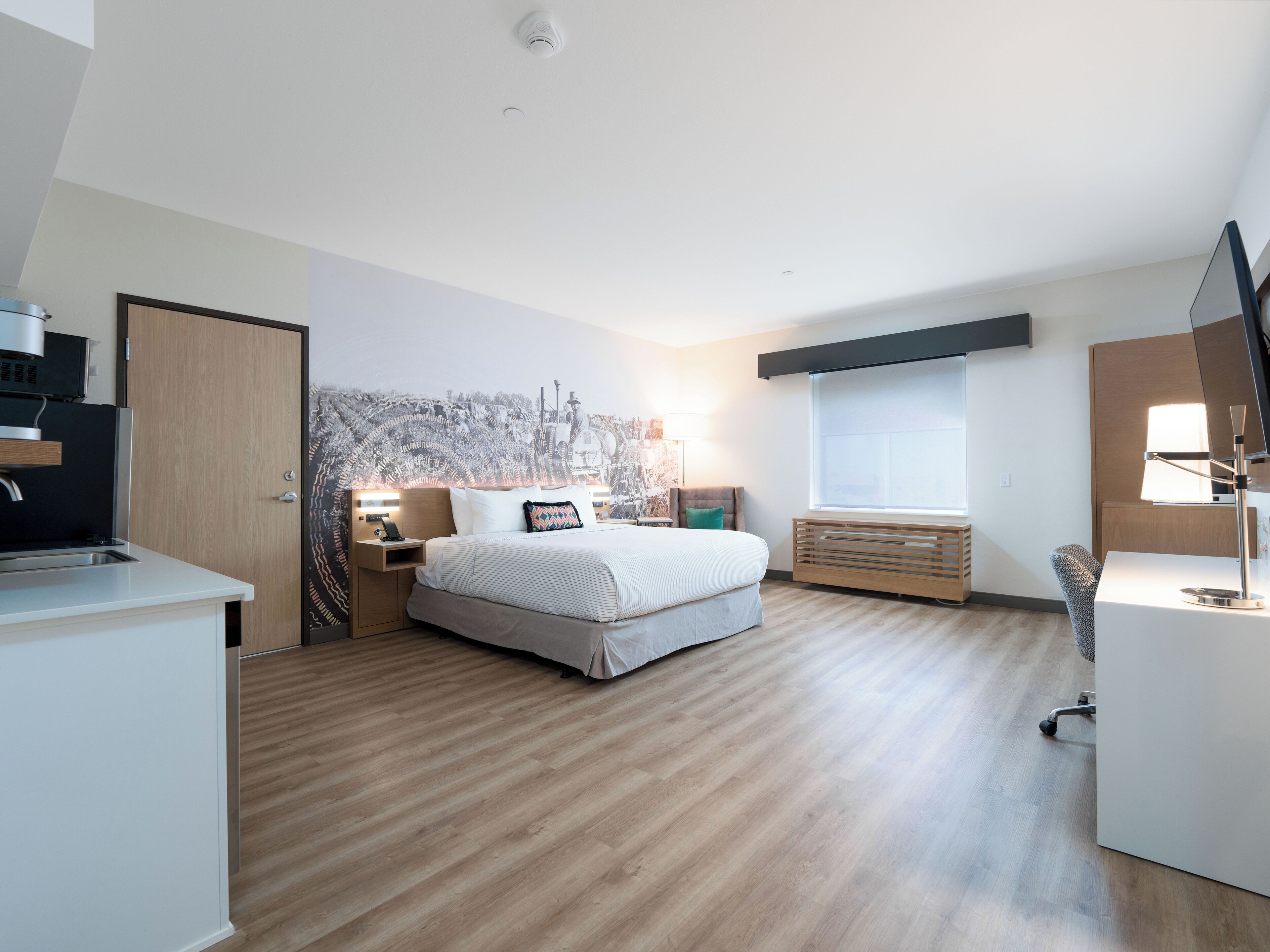 A modern hotel room with a bed, a desk, a chair, a window, and a kitchenette.