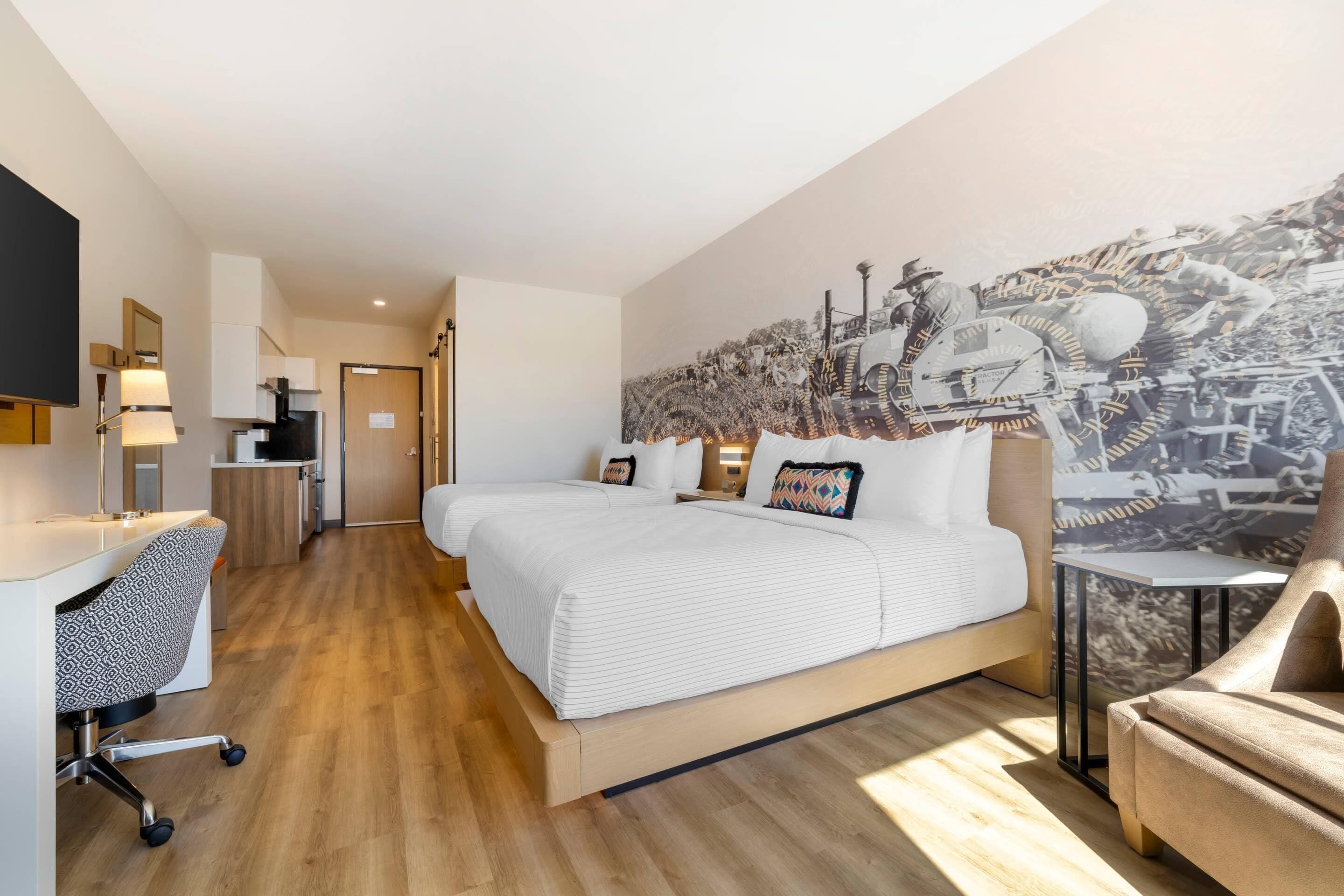 A modern hotel room with a large bed, a desk, and a chair. There is a mural on the wall and a small table next to the bed.