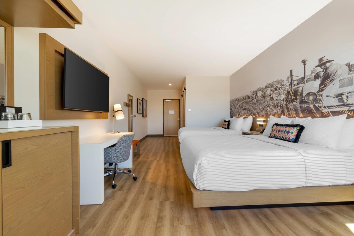 A hotel room with a large bed, a desk with a chair, a flat-screen TV mounted on the wall, and a mural on the wall.
