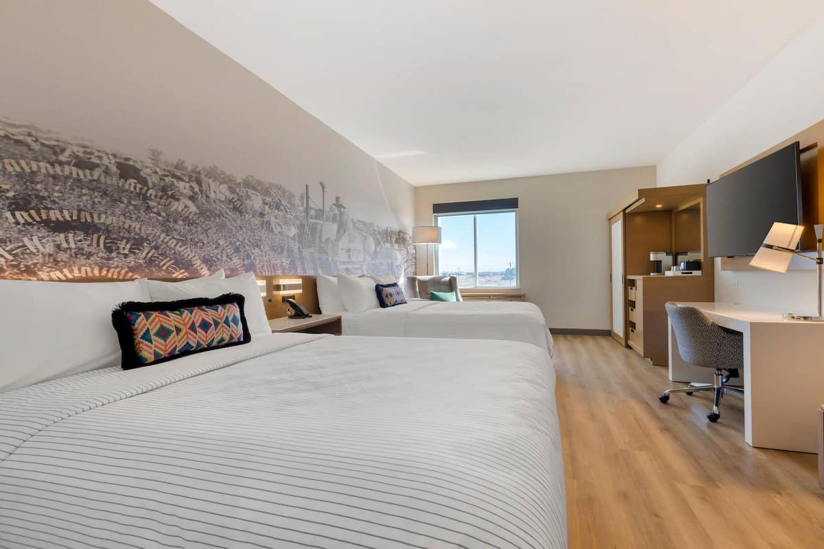 Habitación de hotel con dos camas, un escritorio, una silla, un ventanal grande y televisor montado en la pared.