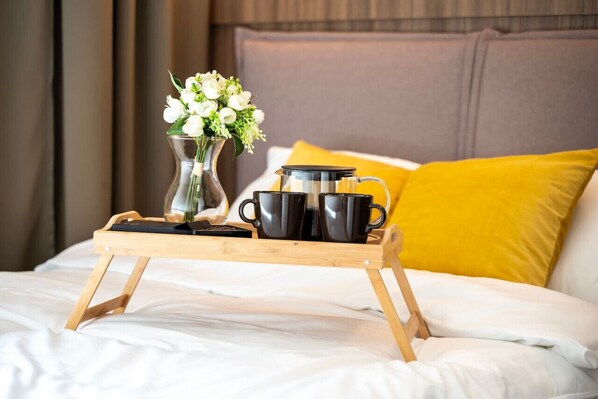 A bed with white bedding, a wooden tray with black mugs and a clear vase with white flowers, and a yellow pillow.