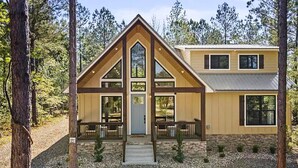 A two-story log cabin with a prominent gable roof, a front porch with benches, and a stone foundation. - It Takes A Village | Poolside Retreat for Groups (Broken Bow)