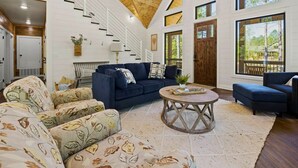 A spacious living room with a dark blue sofa, patterned armchairs, and a round coffee table. A wooden staircase with white railings leads to an upper level. Large windows provide natural light and views of the outdoors. - It Takes A Village | Poolside Retreat for Groups (Broken Bow)