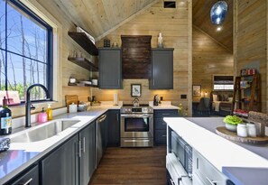 A modern kitchen with stainless steel appliances, wooden cabinets, and a large window providing a view of the outdoors. - It Takes A Village | Poolside Retreat for Groups (Broken Bow)