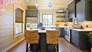 A kitchen with wooden walls, a central island with two wicker chairs, a refrigerator, stove, and a window with blinds. - It Takes A Village | Poolside Retreat for Groups (Broken Bow)