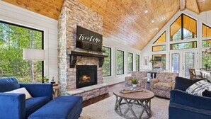 A spacious living room with a stone fireplace, blue sofas, and large windows offering a view of the outdoors. - It Takes A Village | Poolside Retreat for Groups (Broken Bow)