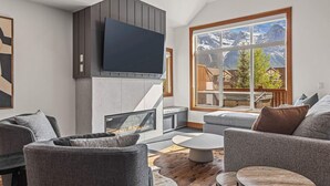 A modern living room with a fireplace, grey sofas, a flat-screen TV, and a large window with a mountain view. - Designer Home, Private Hot Tub (Canmore)