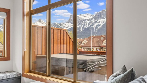 A room with a large window offering a view of snow-capped mountains and traditional houses. - Designer Home, Private Hot Tub (Canmore)