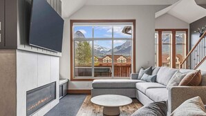 A modern living room with a large window, a fireplace, a grey sofa, and a staircase. - Designer Home, Private Hot Tub (Canmore)