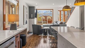 A modern kitchen with a large island, a dining area with a bar, and a view of a mountain house through a large window. - Designer Home, Private Hot Tub (Canmore)