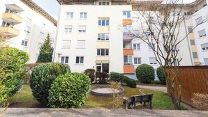 Un edificio residencial de varios pisos con balcones, un pequeño jardín con un banco y un camino de acceso circular.