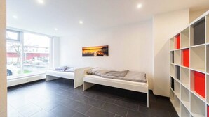 Una habitación moderna con una cama blanca, un ventanal con vistas a edificios y una estantería con secciones rojas y negras.