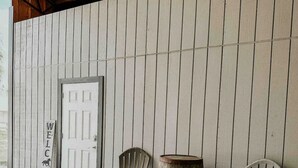 Une véranda couverte avec deux fauteuils Adirondack et un tonneau en bois. On peut voir une pancarte « BIENVENUE » sur le mur.