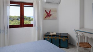 Ett sovrum med en säng, ett fönster med gardiner, en väggmonterad luftkonditioneringsanläggning, ett skrivbord i trä, en stol och en blå kista med en bok i.