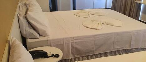 A hotel room with a bed, pillows, a bedside table with a phone, and a kitchenette area with a microwave and cabinets.