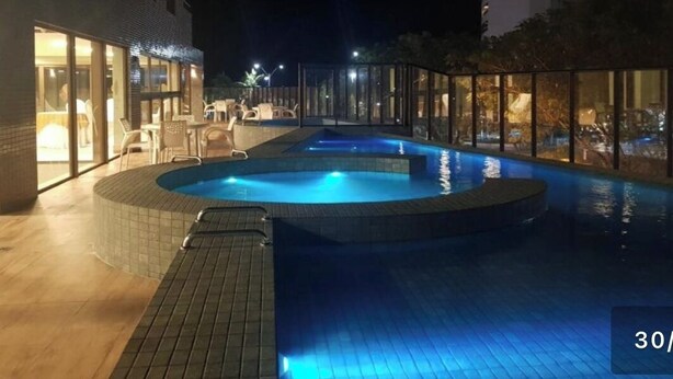 An indoor swimming pool with a tiled edge, surrounded by glass walls and a wooden deck. - ILUSION VISTA PANORÂMICA (Natal)