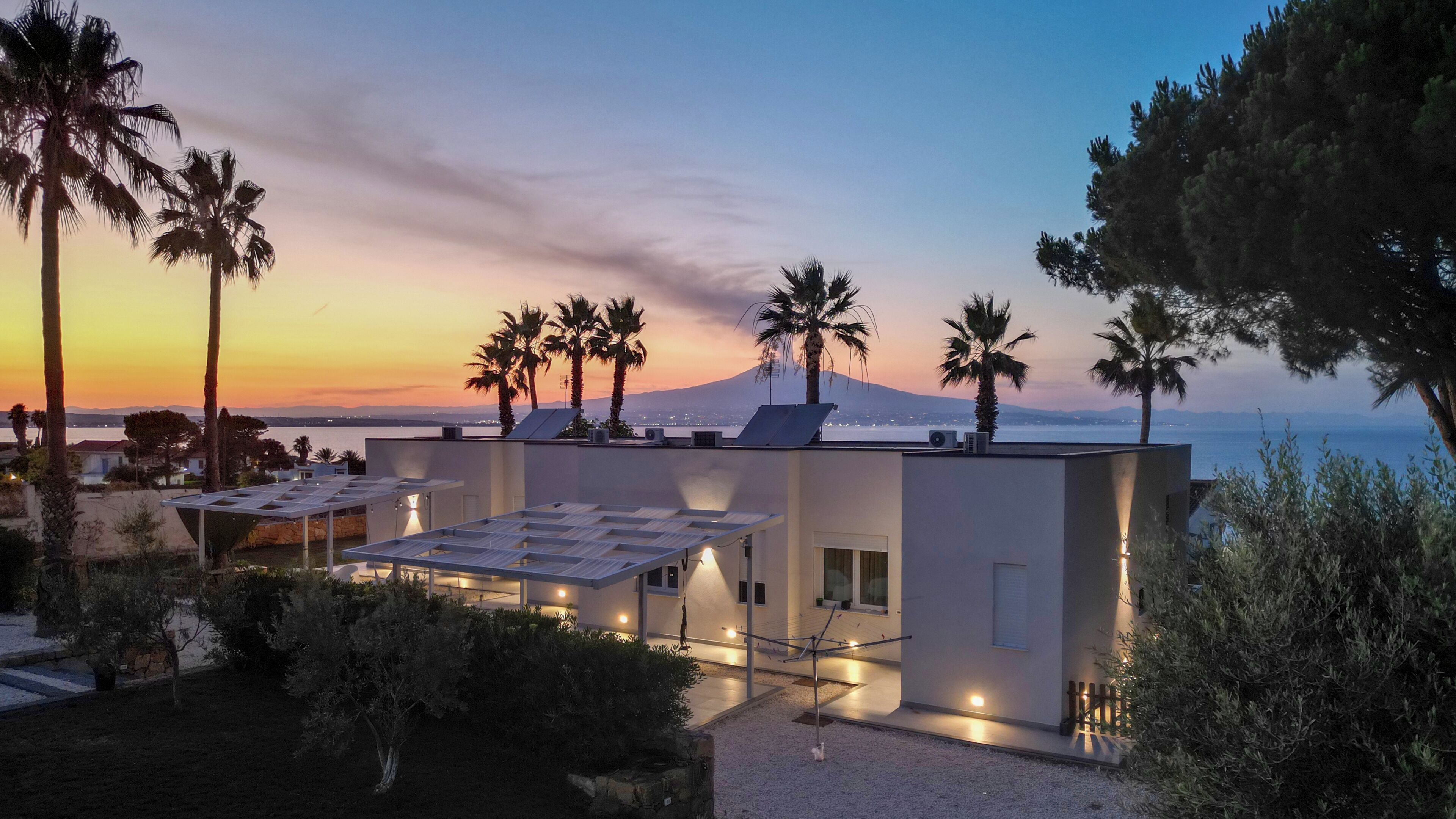 A modern building with a flat roof, solar panels, and a view of the sea and a mountain at sunset.