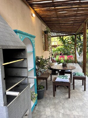 An outdoor patio with a stone barbecue, wooden tables, and chairs, surrounded by potted plants and a wooden pergola. - Pousada Villa dos Tucanos Camburi (Praia de Camburí São Sebastião)