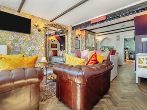 A cozy living room with a brown leather sofa, colorful pillows, and a floral wallpaper. A large mirror reflects the room, and a sign reads "What if the Hokey Cokey really is all about?".