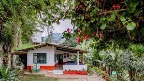 A small, single-story building with a porch, surrounded by lush greenery and flowering plants. - Finca Umnisa  Choachi (Choachí)