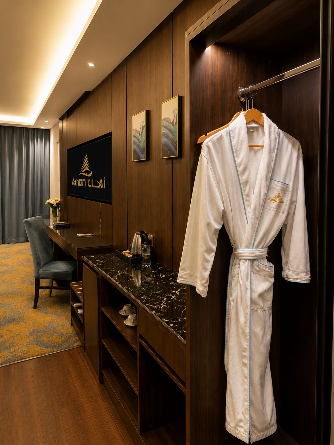 A hotel room with a wooden panel wall, a flat-screen TV, a robe hanging on a rack, a desk with a coffee maker, and a chair.