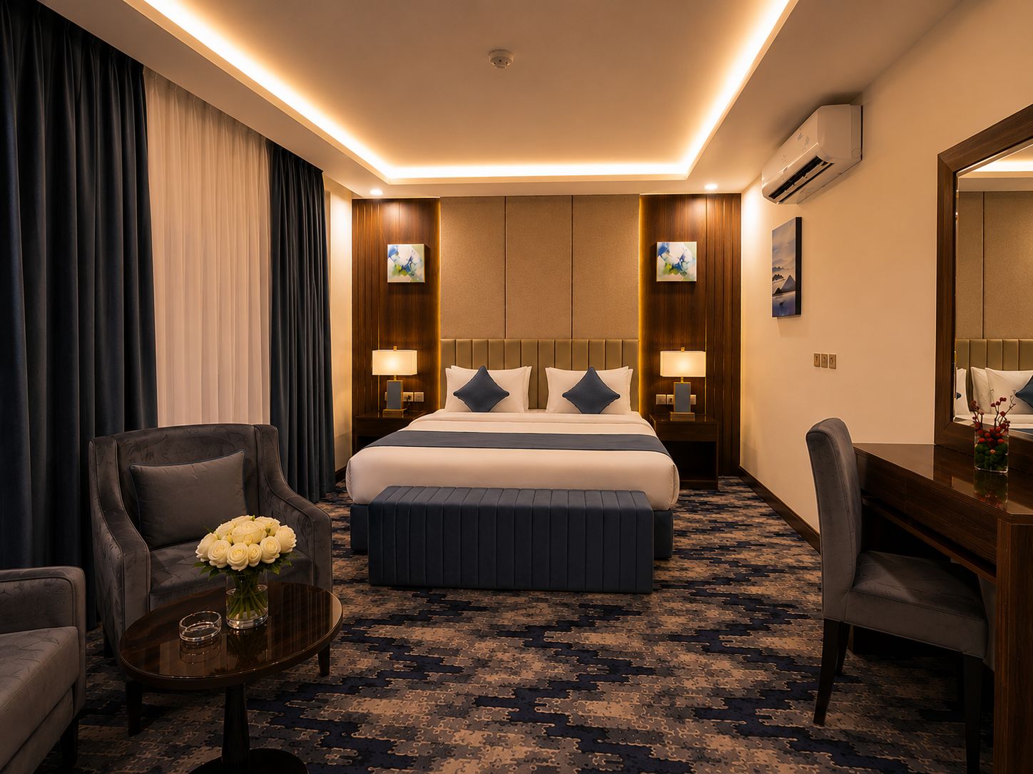 A hotel room with a large bed, two bedside tables with lamps, a desk, a chair, a coffee table with flowers, and a carpeted floor.