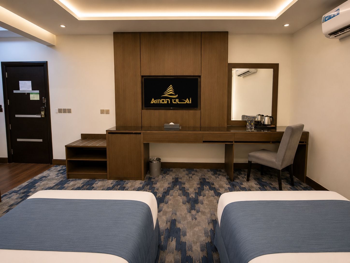 A hotel room with a desk, two beds, a television, and an air conditioning unit.