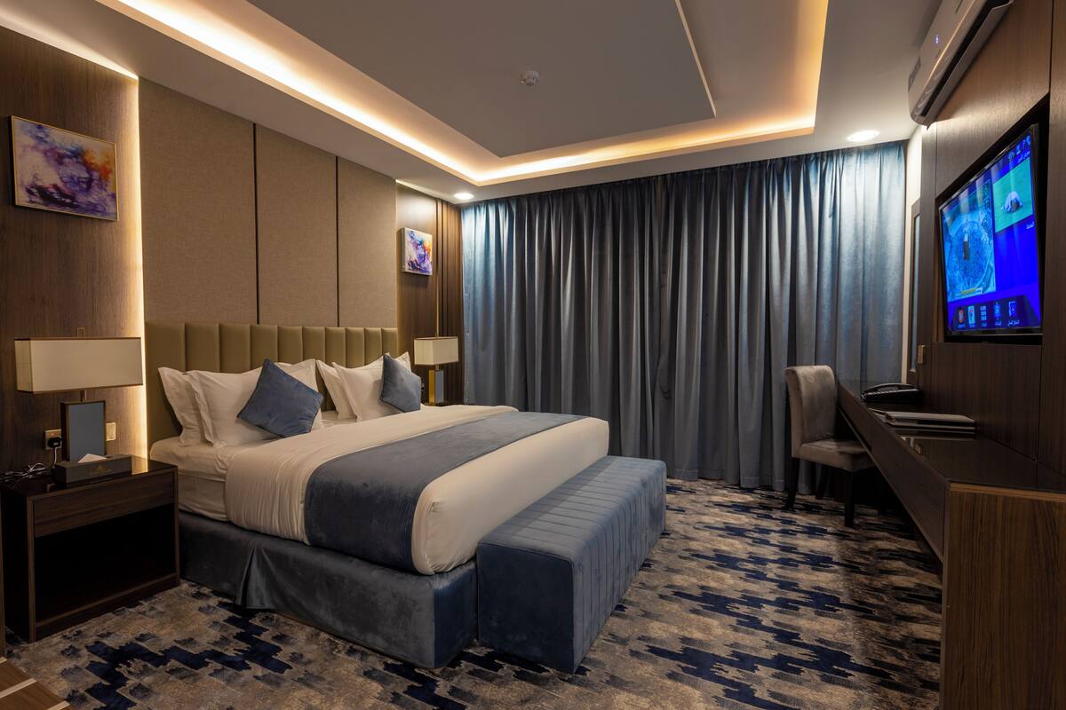 A modern hotel room with a large bed, bedside tables, a desk, a chair, and a flat-screen TV.