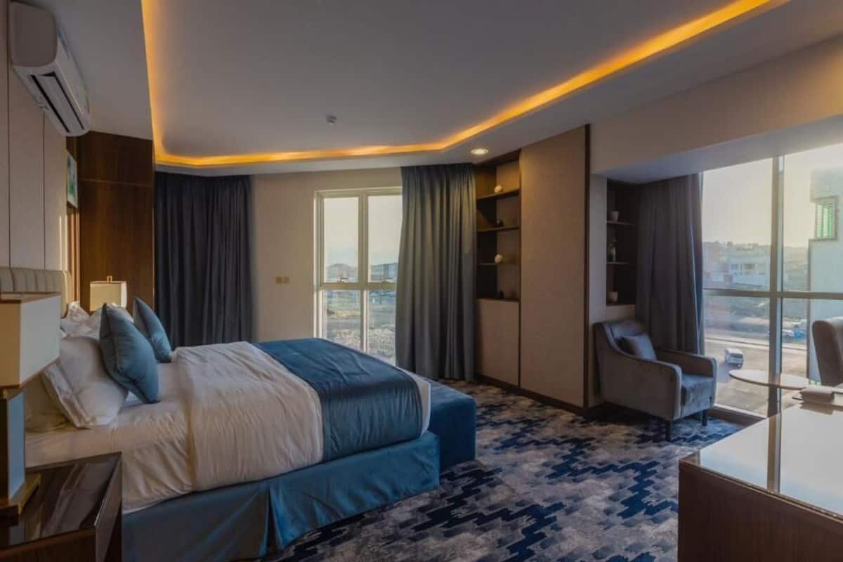 A hotel room with a bed, a desk, a chair, a large window, and a view of the city.