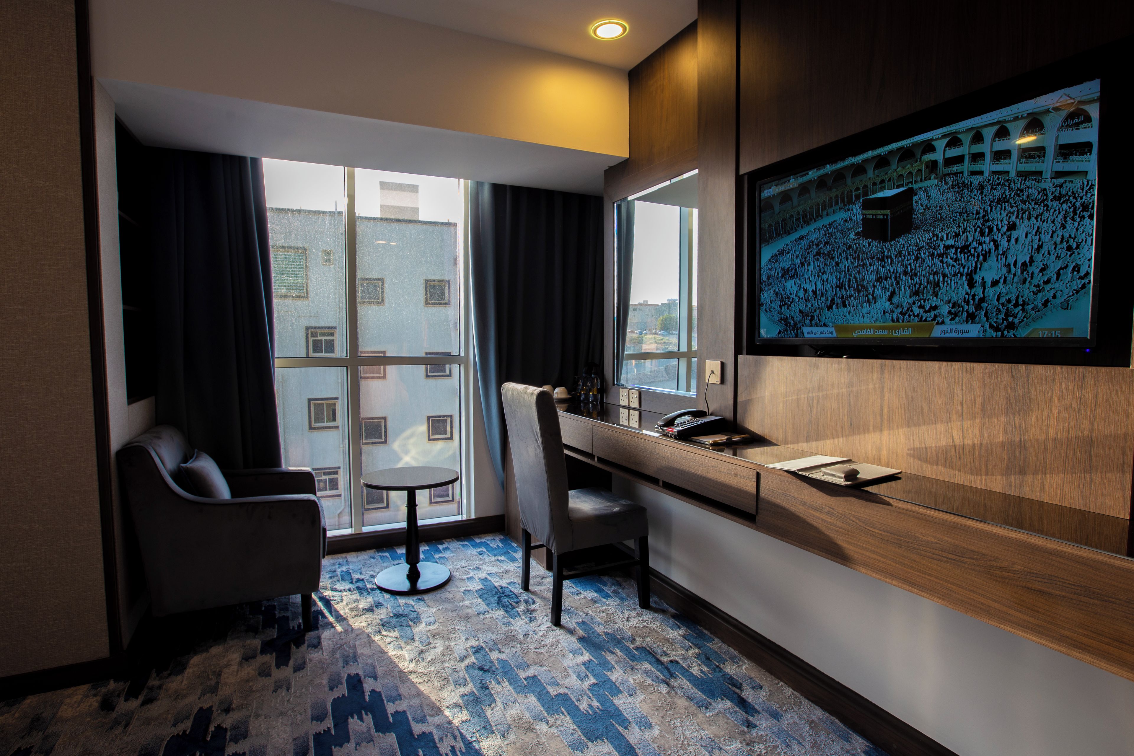 A hotel room with a large window, a desk, a chair, and a television screen displaying a crowd.