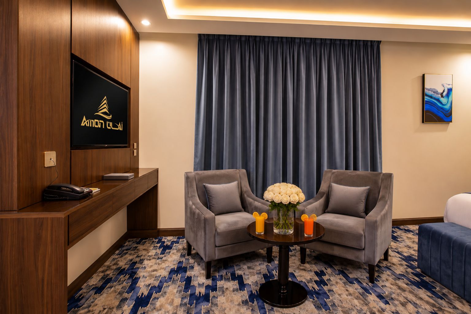 Habitación de hotel con un mostrador de recepción de madera, un televisor de pantalla plana, dos sillones grises, una mesa de centro con un jarrón de flores y una cortina azul.