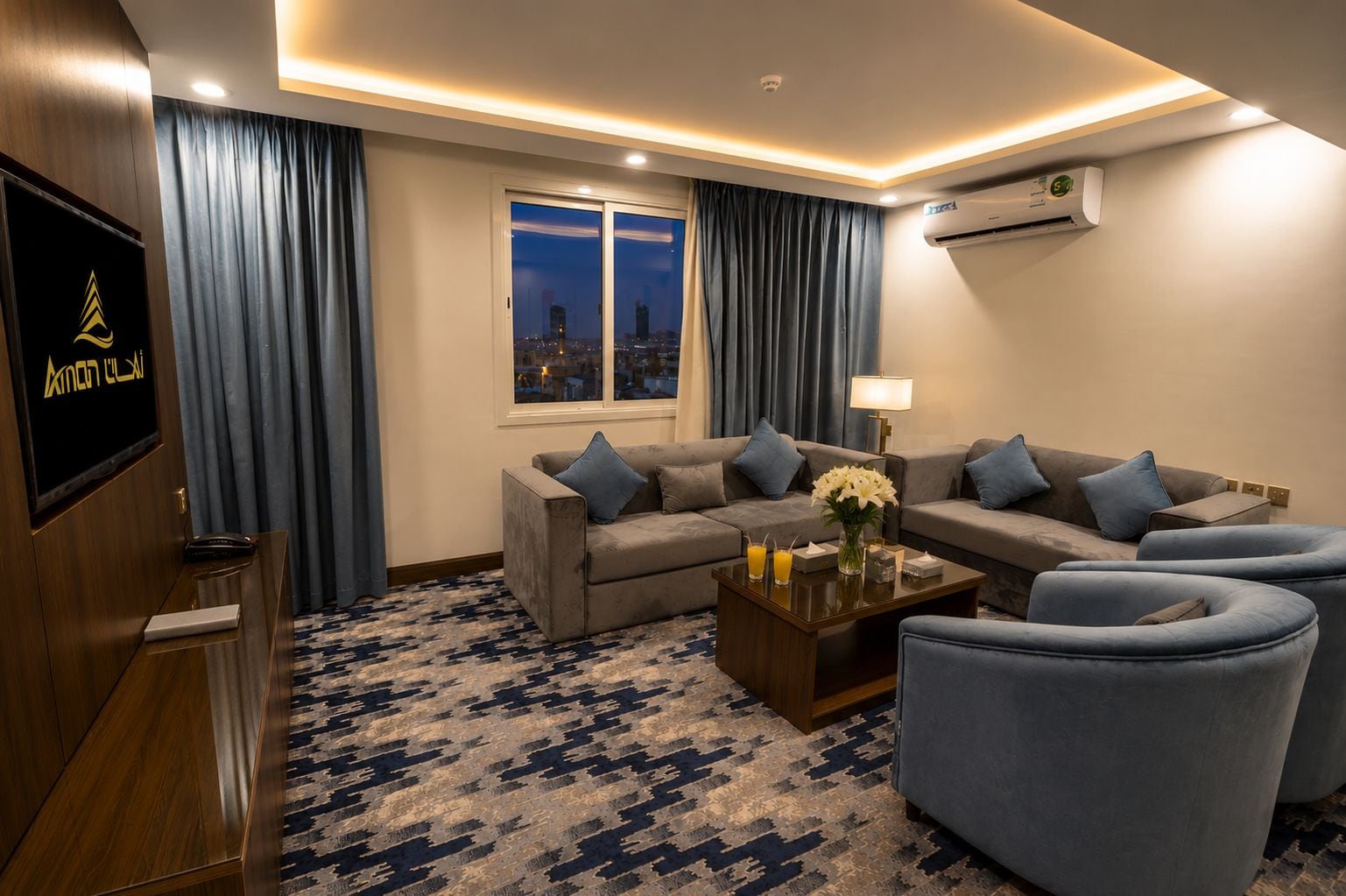Una habitación de hotel moderna con un conjunto de sofás, una mesa de centro y un televisor sobre un mueble de madera.