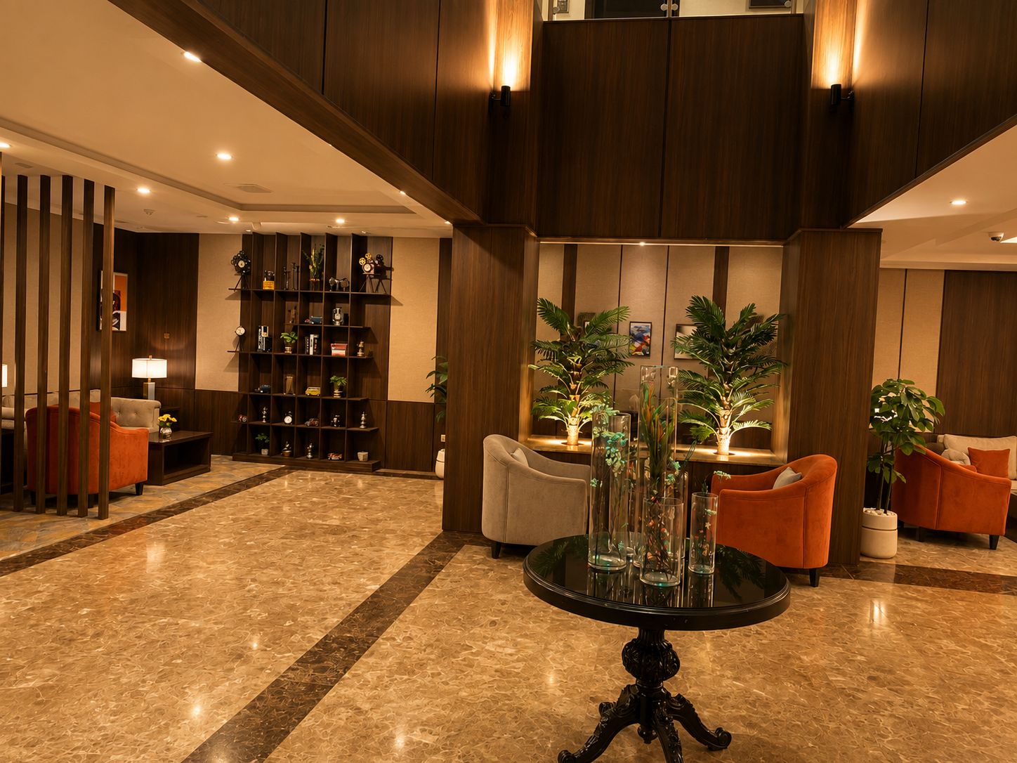 A lobby with a marble floor, a round table with a glass top, orange armchairs, a bookshelf, and two potted plants.