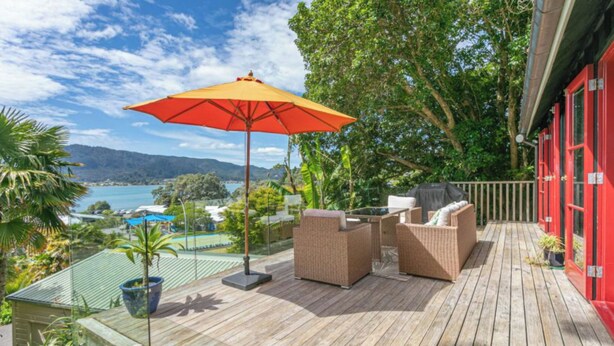 A wooden deck with wicker furniture and a red umbrella, overlooking a scenic bay with mountains in the background.