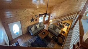 L’intérieur d’un chalet en bois présente un espace de vie avec un ensemble de canapés, une table basse et un ventilateur de plafond. Un escalier avec une rampe en bois est visible au premier plan.