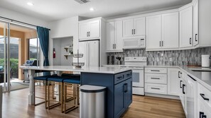 A modern kitchen with white cabinets, a central island with bar stools, a stainless steel trash can, and a view of the outdoors through large windows.