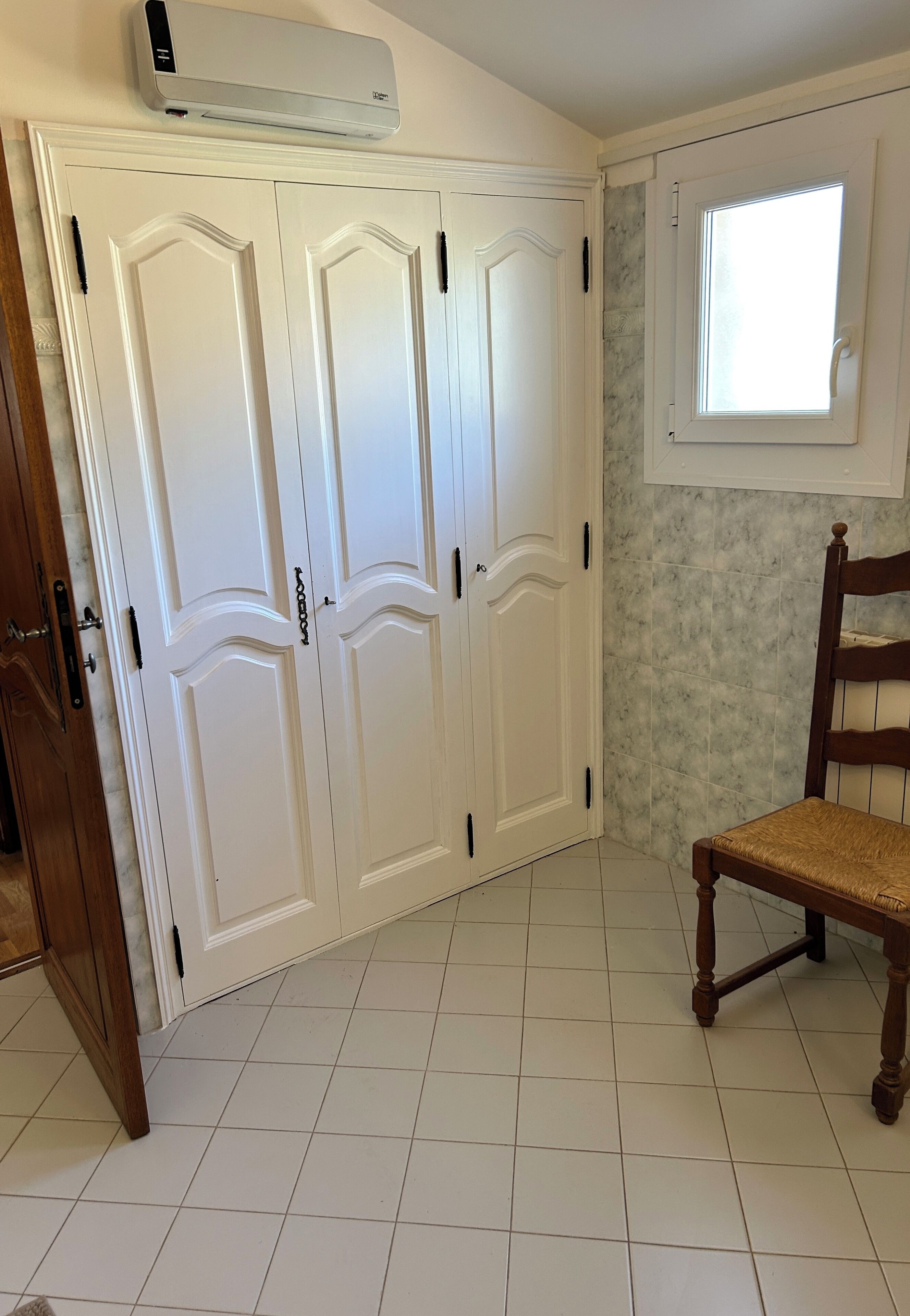 Habitación con suelo de baldosas blancas, una silla de madera, un armario blanco con ventana y un aire acondicionado instalado en la pared.