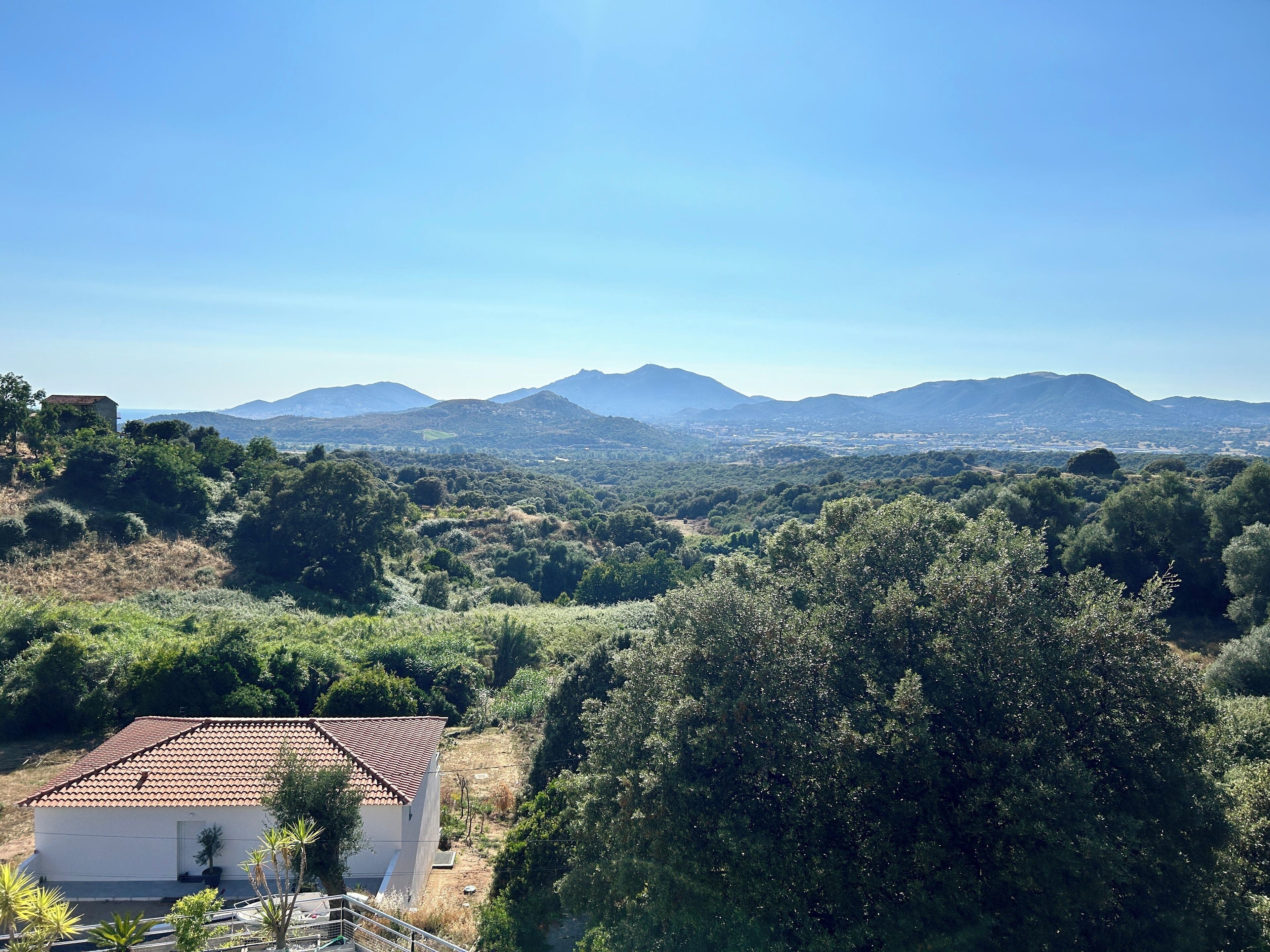 A rural landscape with a white building with a tiled roof, surrounded by dense greenery and mountains in the background.