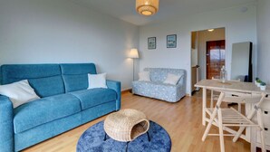 Un salon avec un canapé bleu, un canapé à motifs, un pouf rond en osier, une table à manger en bois avec des chaises et un luminaire suspendu.