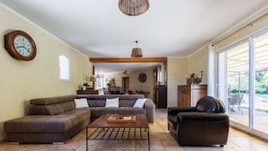 A spacious living room with a sectional sofa, a coffee table, and a leather armchair. A dining area is visible in the background with a kitchen counter and a window. - La Maison de Mathis - Maison avec piscine privée (Valréas)