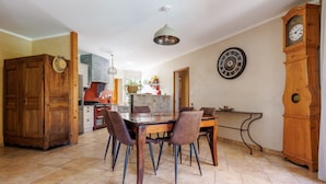 A dining area with a wooden table and chairs, a kitchen in the background, and a large wall clock. - La Maison de Mathis - Maison avec piscine privée (Valréas)