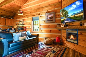 A cozy cabin interior with a leather sofa, a fireplace, wooden walls, and a television displaying a scenic view. - Point of View Cabin · Private Hot Tub, Fire Pit & Mountain Views · Dog-Friendly (Sevierville)
