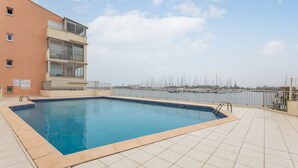 Una piscina con vistas a veleros a lo lejos, rodeada por una solana con baldosas y un edificio con balcones.